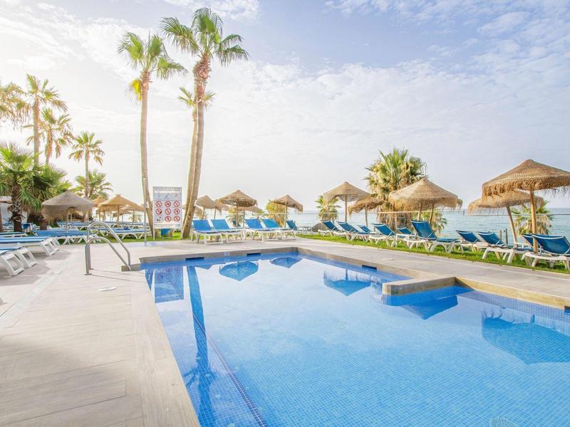 Schwimmbad mit blauer Wasserfläche, Liegen, Sonnenschirmen und Palmen bei sonnigem Wetter.