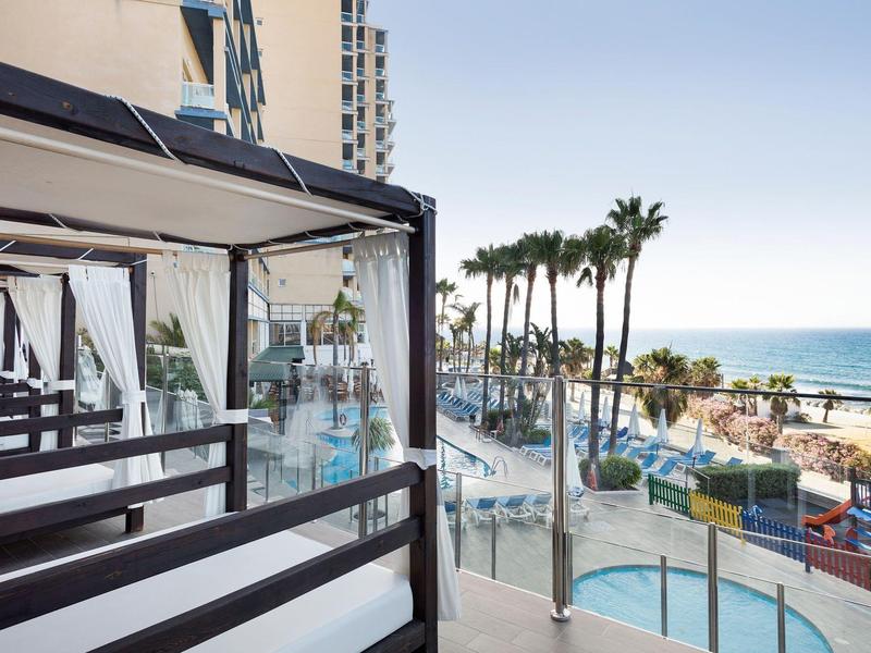 Balkon mit schwarzem Geländer, Blick auf Pool, Palmen und das blaue Meer im Hintergrund.