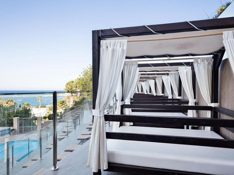 Weiße Überdachungen mit Vorhängen am Pool, Meerblick im Hintergrund, klarer Himmel, helle Farben.
