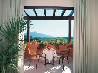 Vue d'une chambre sur une terrasse avec table et chaises, plantes et paysage montagneux en arrière-plan.