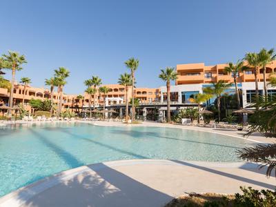 Grand piscine extérieure devant un bâtiment d'hôtel à plusieurs étages avec des palmiers sous un ciel bleu clair.