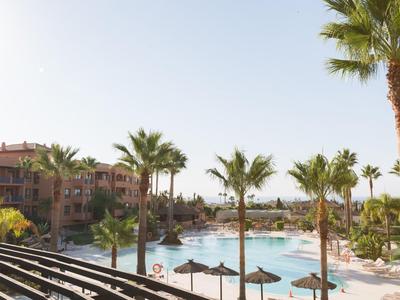 Großer Hotelpool umgeben von Palmen und Sonnenschirmen bei klarem Himmel.