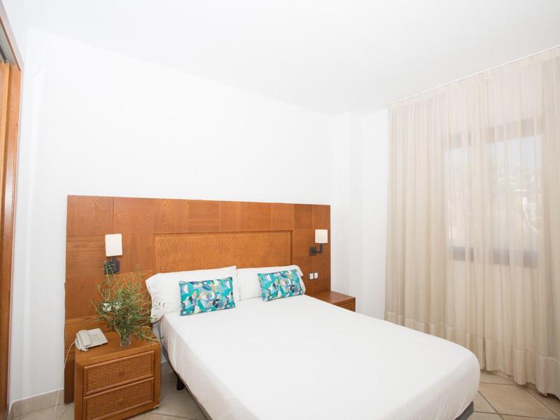 Helles Hotelzimmer mit Doppelbett, Holzkopfteil und Nachttisch mit Pflanze und Telefon.