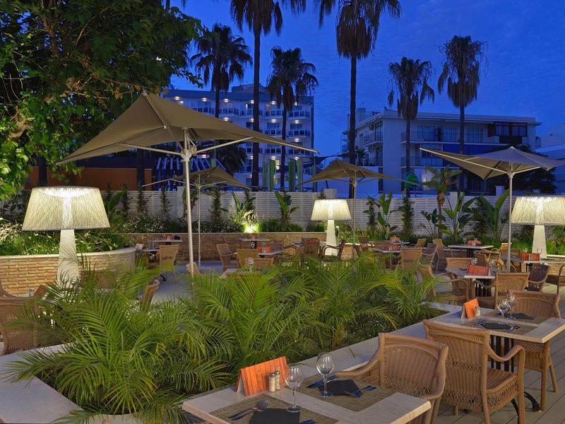 Abendliche Terrasse mit Tischen, Stühlen, Lampen und Sonnenschirmen vor Palmen und Gebäuden.