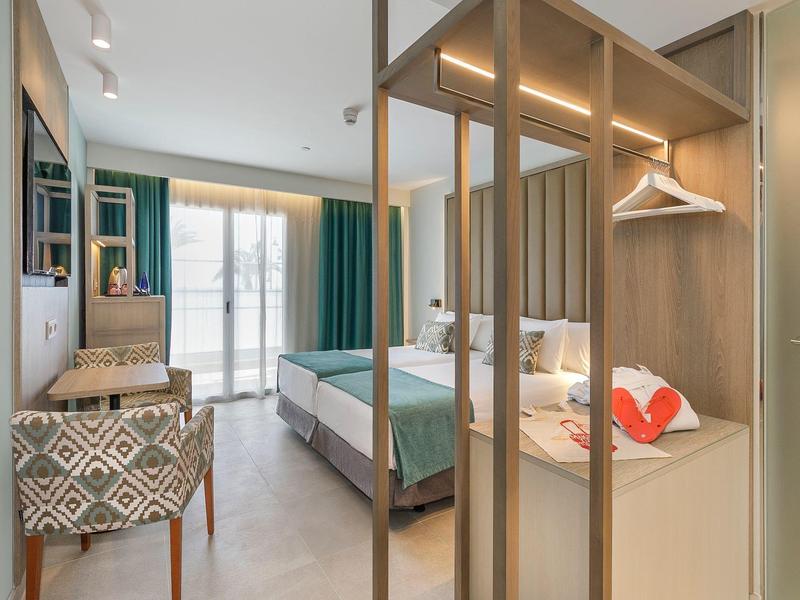 Helles Hotelzimmer mit Doppelbett, grün-blauen Vorhängen und offener Garderobe aus Holz und Metall.