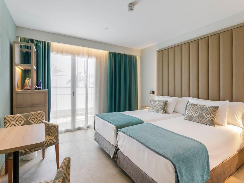 Helles Zimmer mit zwei Betten, türkisfarbenen Vorhängen, Holzmöbeln und großem Fenster.