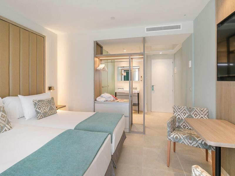 Modernes Hotelzimmer mit zwei Betten, gepolstertem Kopfteil, Glaswand und kleinem Tisch mit Stühlen.