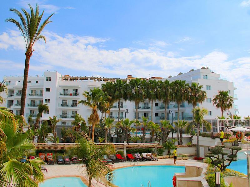Großer Pool mit Palmen und weißem Hotelgebäude im sonnigen Tropenresort unter blauem Himmel.