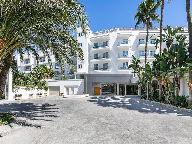 Weißes Hotelgebäude mit Balkonen, tropischen Palmen und großzügigem Eingangsbereich bei Sonnenschein.