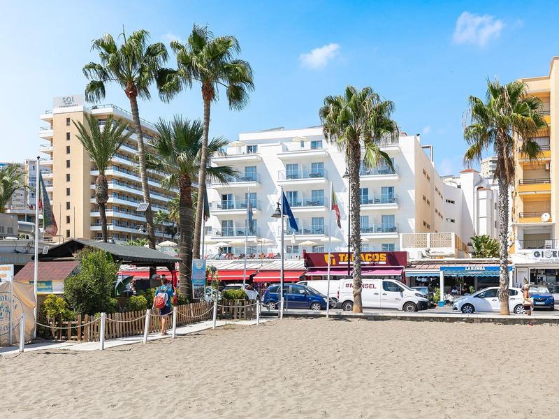 Sandstrand mit Palmen und mehrstöckigen Wohngebäuden im Hintergrund bei blauem Himmel.