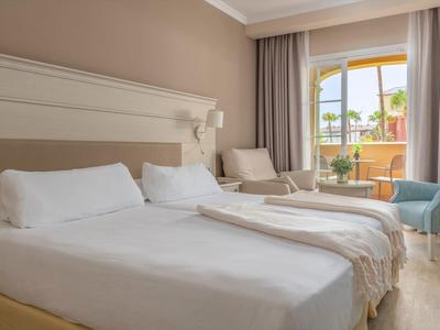 Gemütliches Hotelzimmer mit zwei Einzelbetten, Sessel, Schreibtisch und Balkon mit Blick nach draußen.