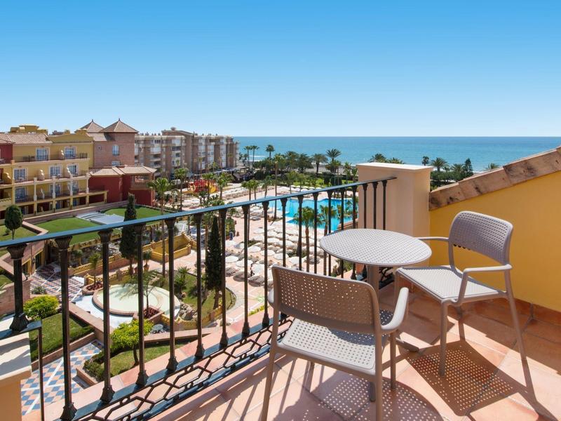 Balkon mit Tisch und Stühlen, Blick auf Pool, Palmen, Häuser und blauen Himmel am Meer.