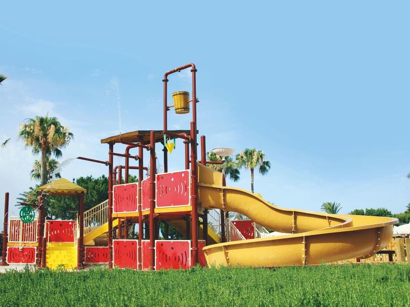 Bunter Spielplatz mit Rutschen und Klettergerüst, umgeben von Gras und Palmen unter blauem Himmel.