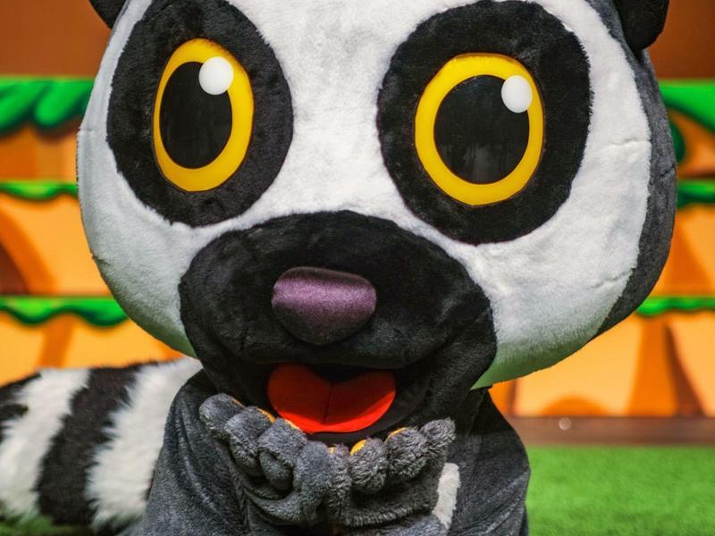 Große graue Tierfigur mit gelben Augen liegt auf grünem Boden vor bunter Wand.