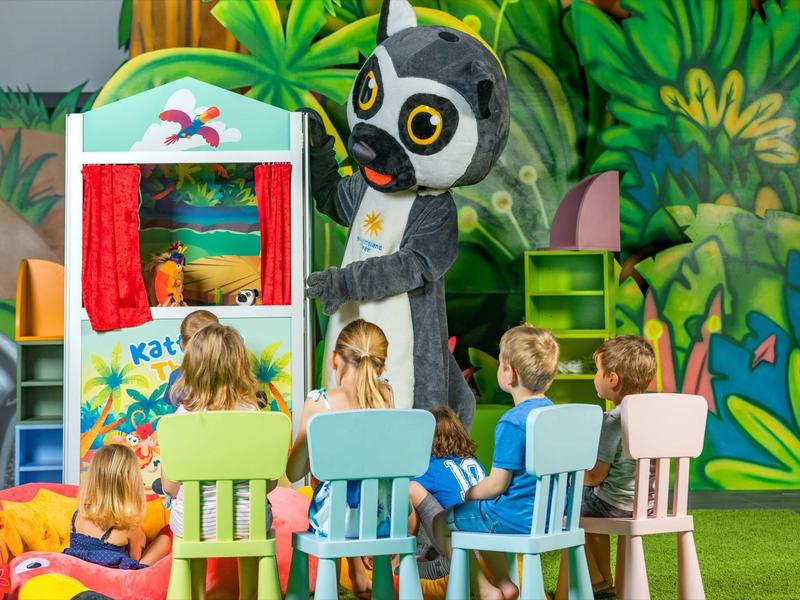 Kinder sitzen auf bunten Stühlen vor einem Kasperletheater, im Hintergrund eine Waldkulisse und eine Tierfigur.