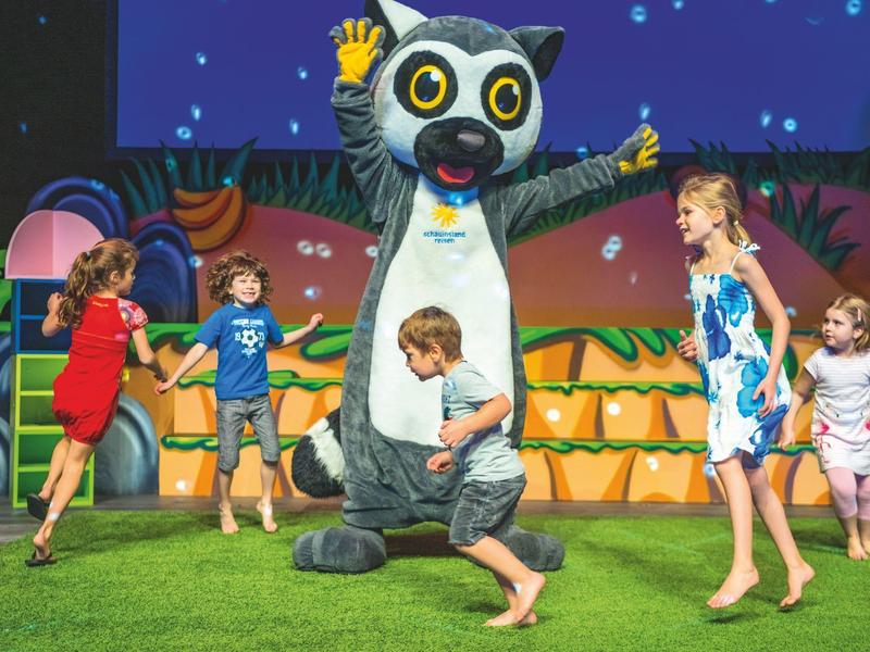 Kinder spielen und tanzen fröhlich um eine große, bunte Tierfigur auf einer Bühne mit grüner Wiese.