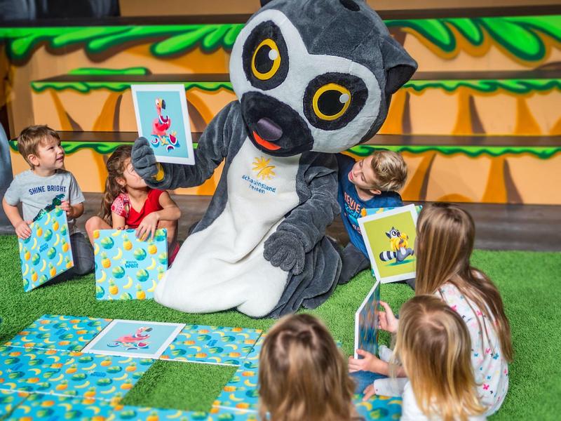 Große Eule mit Kindern auf grünem Teppich, bunte Bilder haltend, bunte Umgebung, fröhliche Szene.