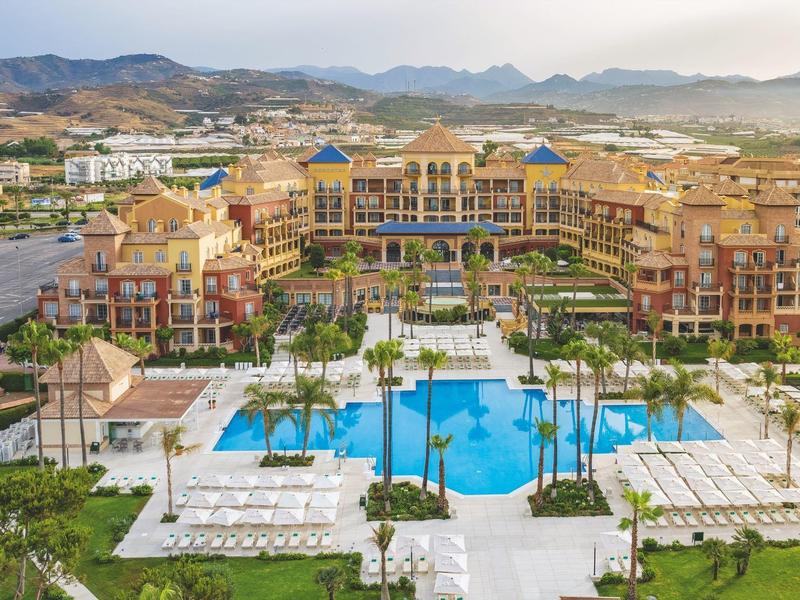 Großes Resort mit blauem Pool, umgeben von Palmen und Bergen im Hintergrund.