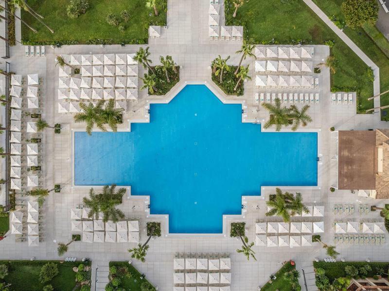 Vogelperspektive auf ein großes, kreuzförmiges Schwimmbecken mit weißen Liegestühlen und grüner Umgebung.