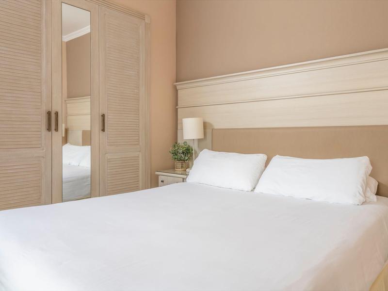 Schlichtes Schlafzimmer mit weißem Bett, Kissen, Nachttisch und hellem Holzkleiderschrank mit Spiegel.