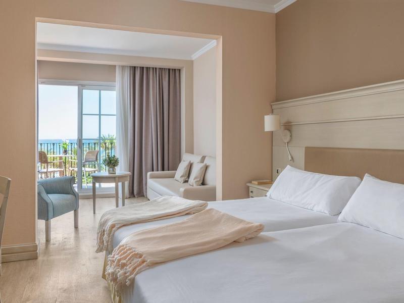 Helles Hotelzimmer mit Doppelbett, Sitzbereich, Balkon und Blick auf das Meer.