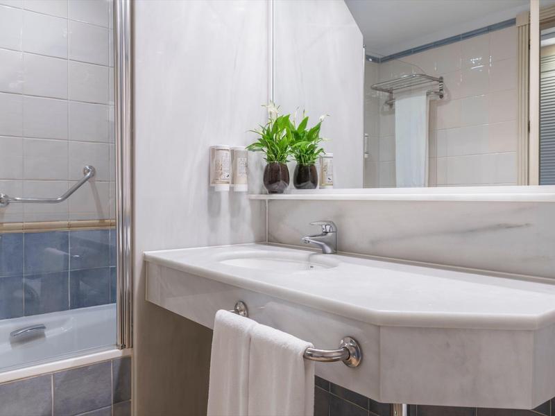 Hell gestaltetes Badezimmer mit großem Spiegel, weißem Waschbecken, Handtüchern und grünen Pflanzen.