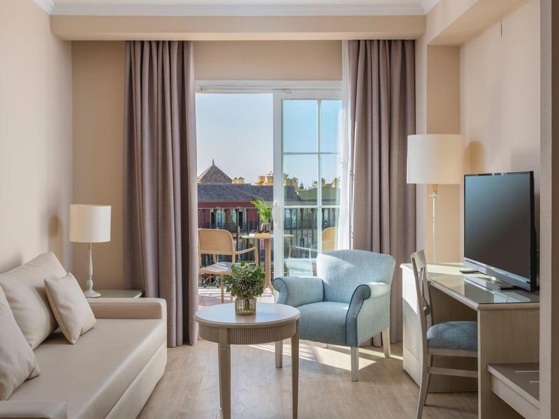 Helles Wohnzimmer mit Sofa, blauem Sessel, kleinem Tisch, TV und Blick auf ein Stadtgebäude.