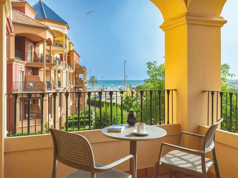 Balkon mit Tisch und Stühlen, Blick auf Meer, Sonne, mediterrane Häuser, grüne Pflanzen, blaue Himmel.