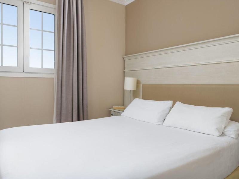 Kleines helles Schlafzimmer mit großem Bett, weißer Bettwäsche, Fenster und grauer Gardine.