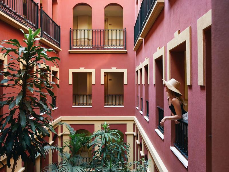 Innenhof mit roten Wänden, mehreren Fenstern und einer sitzenden Frau mit Hut an einem Fenster.