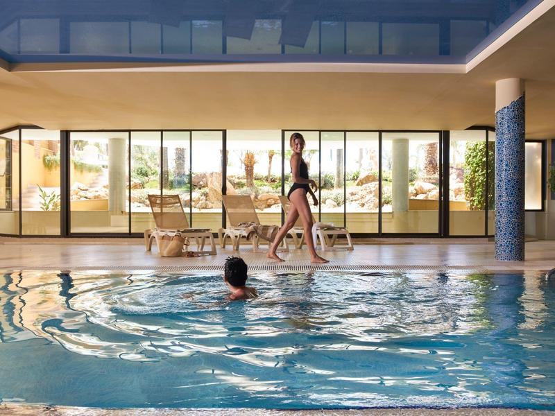 Innenbereich mit großem Swimmingpool, eine Frau läuft am Beckenrand, ein Mann schwimmt im Wasser.