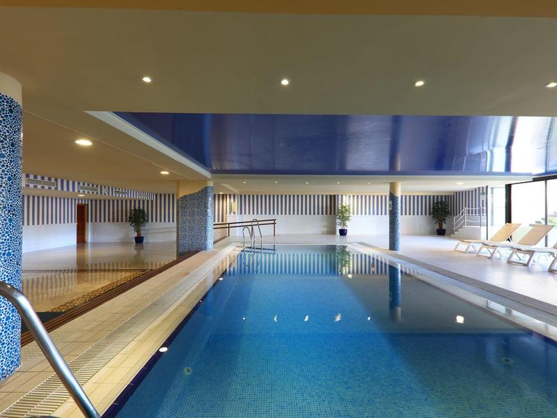 Ein moderner Innenpool mit blauen Fliesen, Holzbereich und weißen Liegestühlen am Fenster.