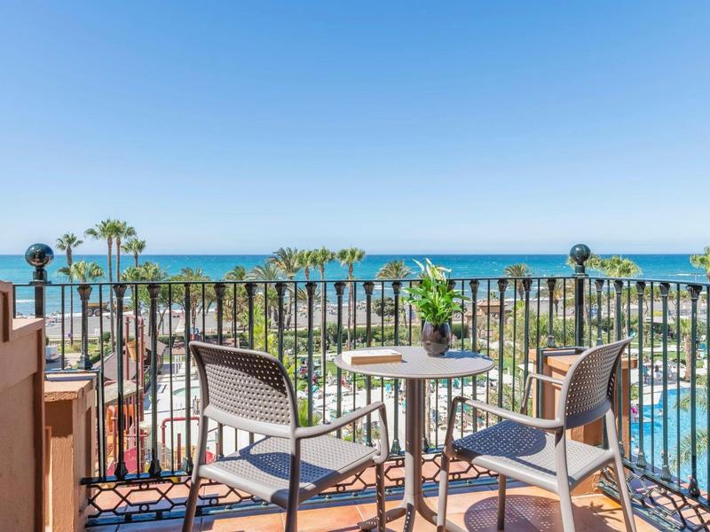 Balkon mit zwei Stühlen und Tisch, Blick auf Palmen, Pool und das blaue Meer im Hintergrund.