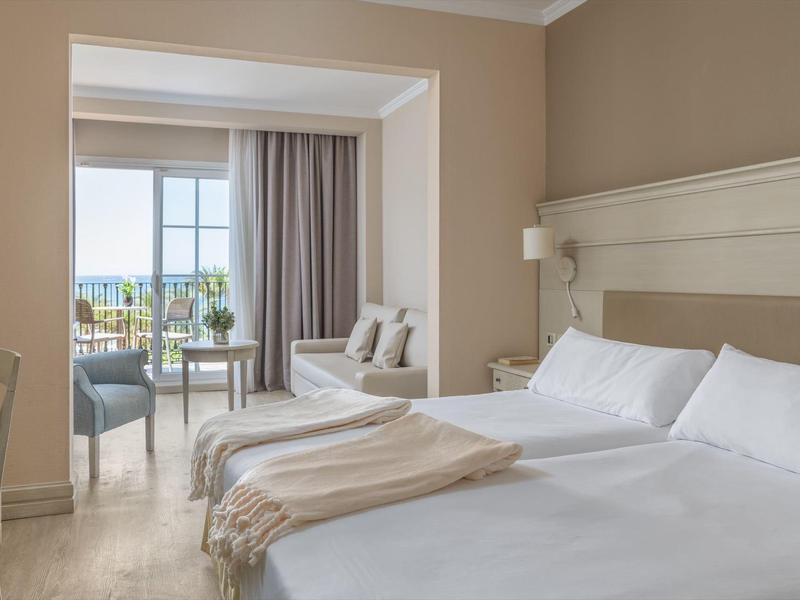 Helles Hotelzimmer mit Doppelbett, Sitzbereich, Balkon und Meerblick bei Tageslicht.