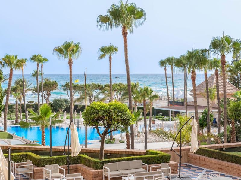 Hotelpool mit Sonnenliegen, Palmen und Blick auf den türkisfarbenen Ozean bei klarem Himmel.