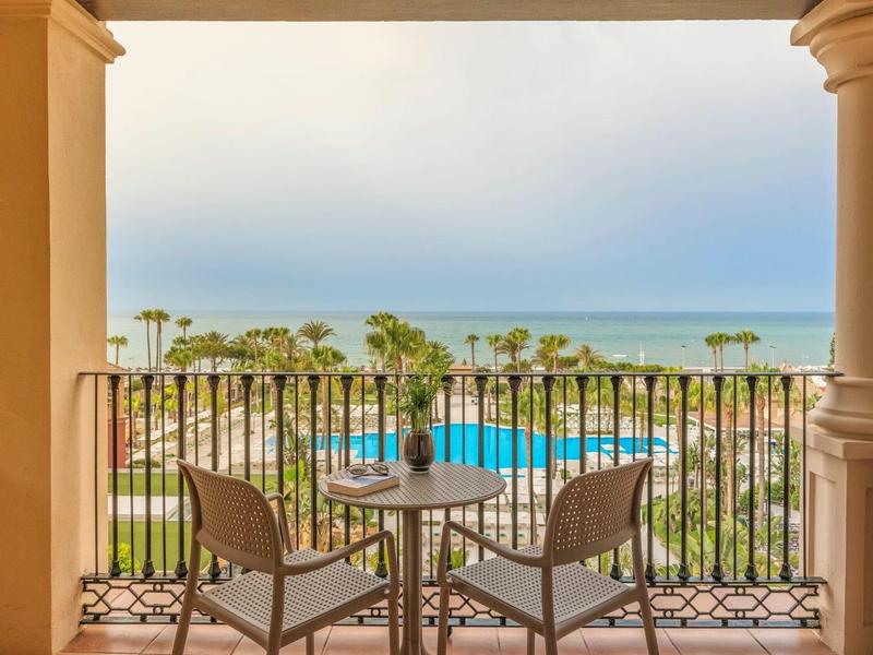 Balkon mit zwei Stühlen und Tisch, Blick auf Palmen, Strand und blauen Himmel am Meer.