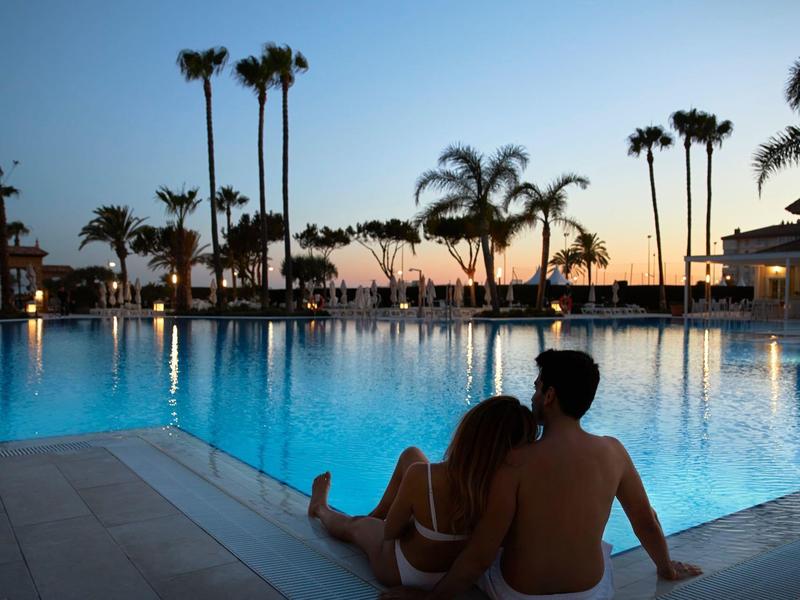 Paar sitzt am Poolrand, Palmen im Hintergrund, Sonnenuntergang, ruhige Atmosphäre, warmes Licht.