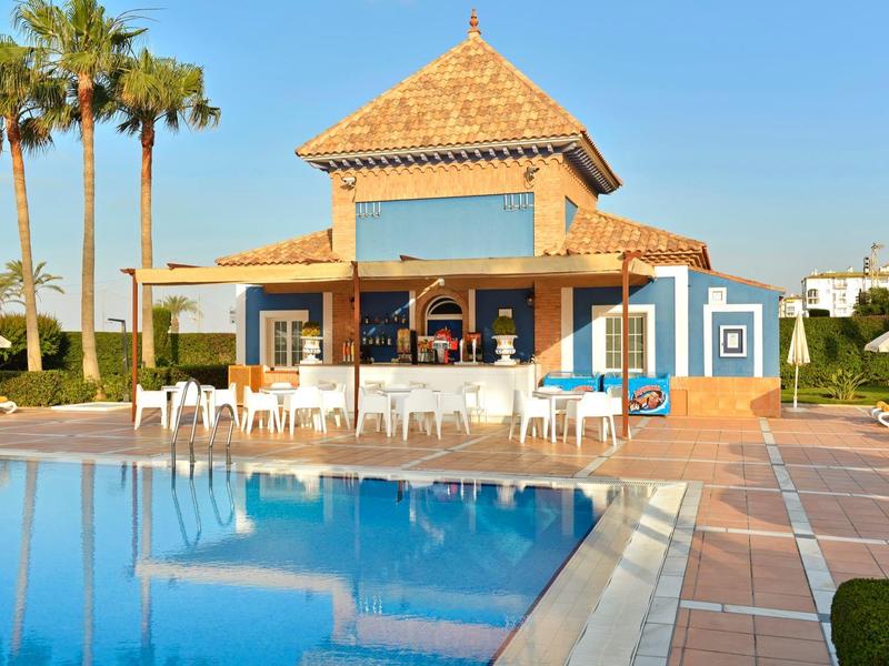 Blauer Pool vor einem kleinen, blau-weißen Poolhaus mit Palmen und klarem Himmel.