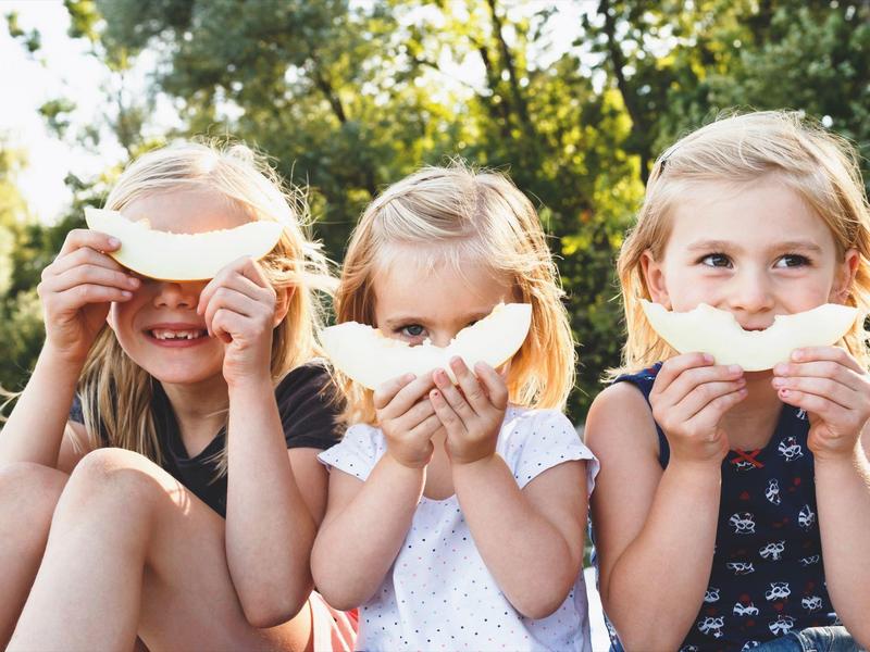 Drei Mädchen sitzen draußen und halten Melonenscheiben vor ihr Gesicht.