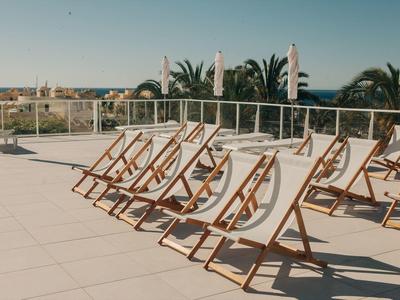 Leere Liegestühle aus Holz mit weißen Planen auf heller Terrasse bei klarem Himmel und Palmen.