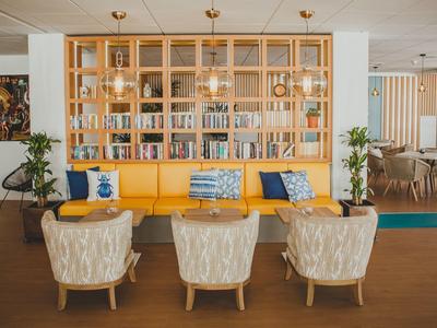 Gemütlicher Wartebereich mit gelber Bank, drei beigen Sesseln, Holzregal und hängender Beleuchtung.