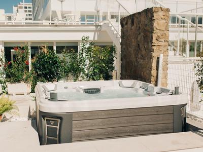 Moderner Whirlpool mit brauner Verkleidung steht im hellen Gartenbereich, umgeben von Pflanzen.