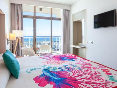 Helles Schlafzimmer mit Meerblick, großem Bett, buntem Bettbezug und Balkon mit Sitzgelegenheiten.