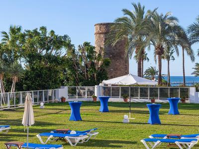 Césped verde con tumbonas, mesas altas azules y palmeras con vista al mar.
