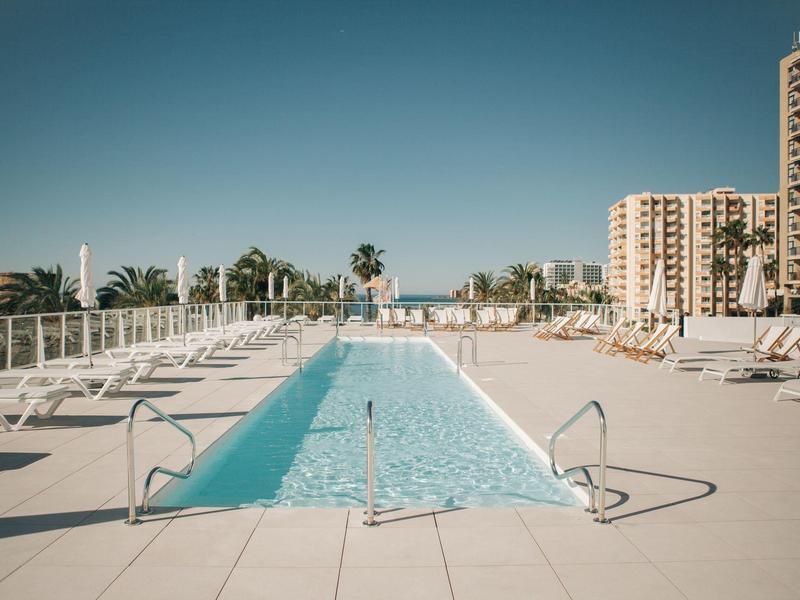 Schmaler rechteckiger Pool mit weißen Liegestühlen und hohen Gebäuden im Hintergrund unter blauem Himmel.
