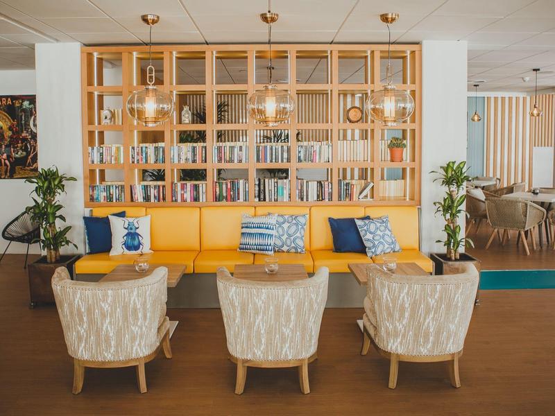 Gemütlicher Wartebereich mit gelber Bank, drei beigen Sesseln, Holzregal und hängender Beleuchtung.