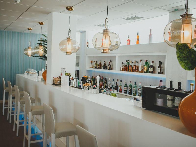 Moderne Bar mit weißen Hockern, großen Glühlampen und Regalen voller bunter Flaschen.