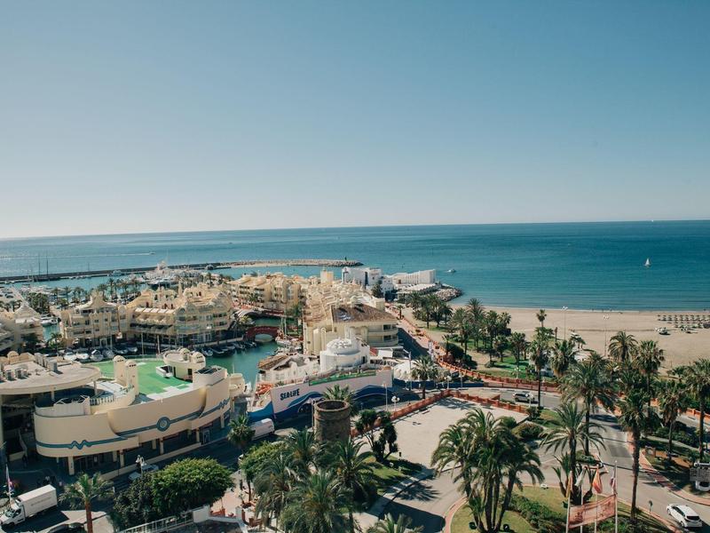 Küstenlandschaft mit Strand, Palmen, blauer See und verschiedenen Gebäuden im Vordergrund.