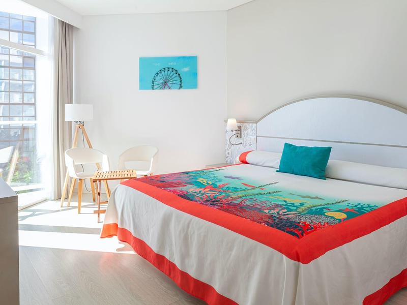 Hell eingerichtetes Schlafzimmer mit großem Fenster, weißem Bett, bunten Decken und blauem Kissen.