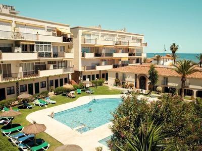 Schwimmbad mit Sonnenliegen, umgeben von weißen Wohngebäuden und Palmen unter blauem Himmel.